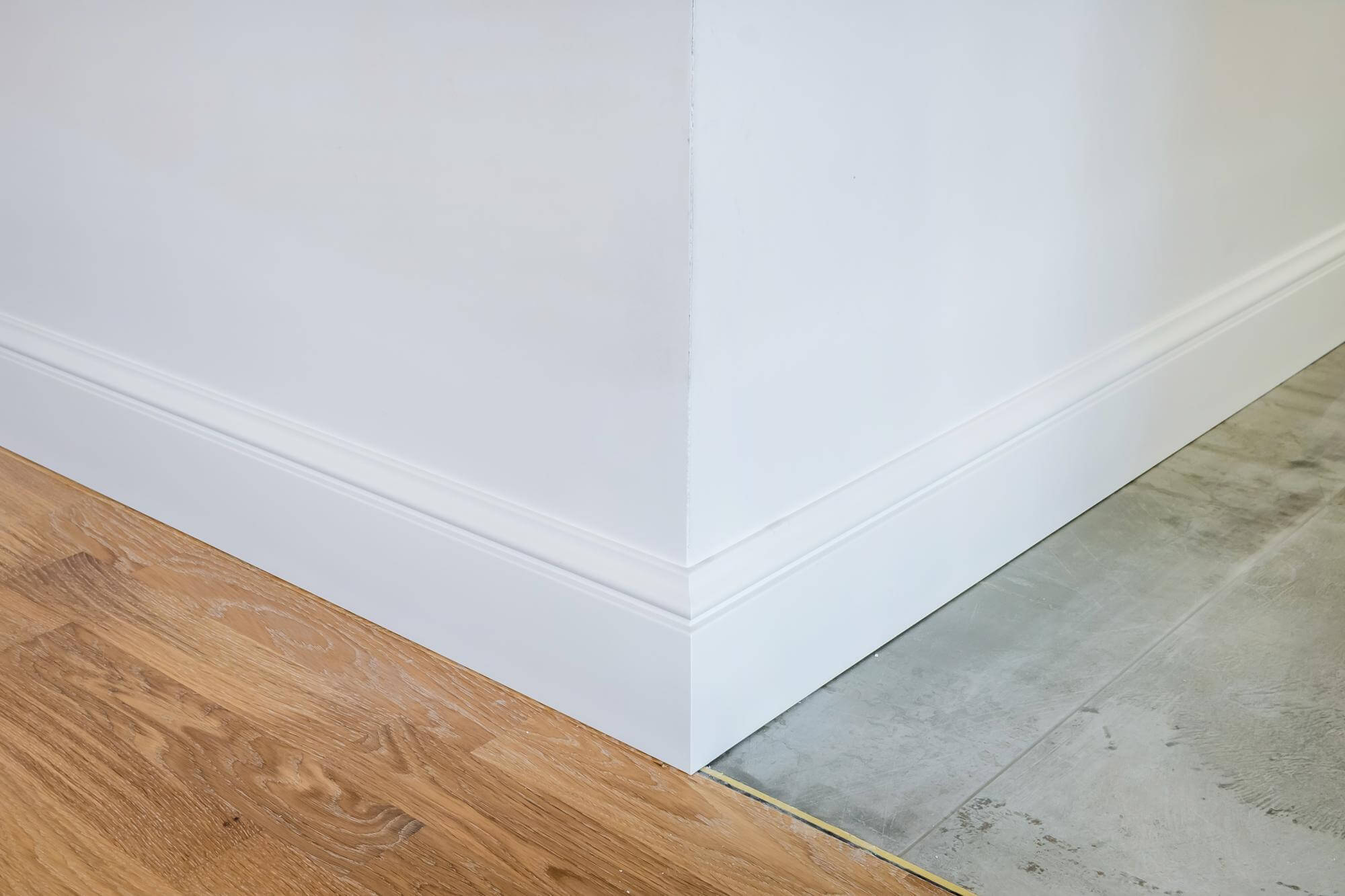 Kulma valkoisella jalkalistalla, jossa on puulattia ja harmaa lattia, valaistus on kirkas.