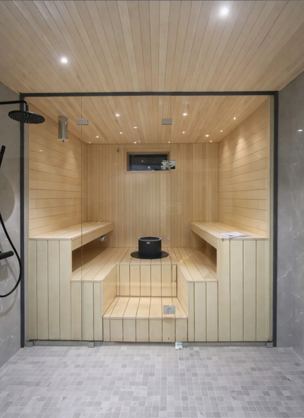 Sauna, jossa puupanelointi, penkit ja kiuas. Harmaa laattalattia ja suihku.